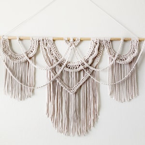 May include: A white macrame wall hanging with a wooden dowel rod and fringe. The hanging features a geometric design with a central diamond shape.