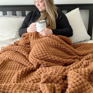 May include: A person is sitting on a bed with a brown waffle knit blanket. They are holding a white mug with the word "miracle" on it.