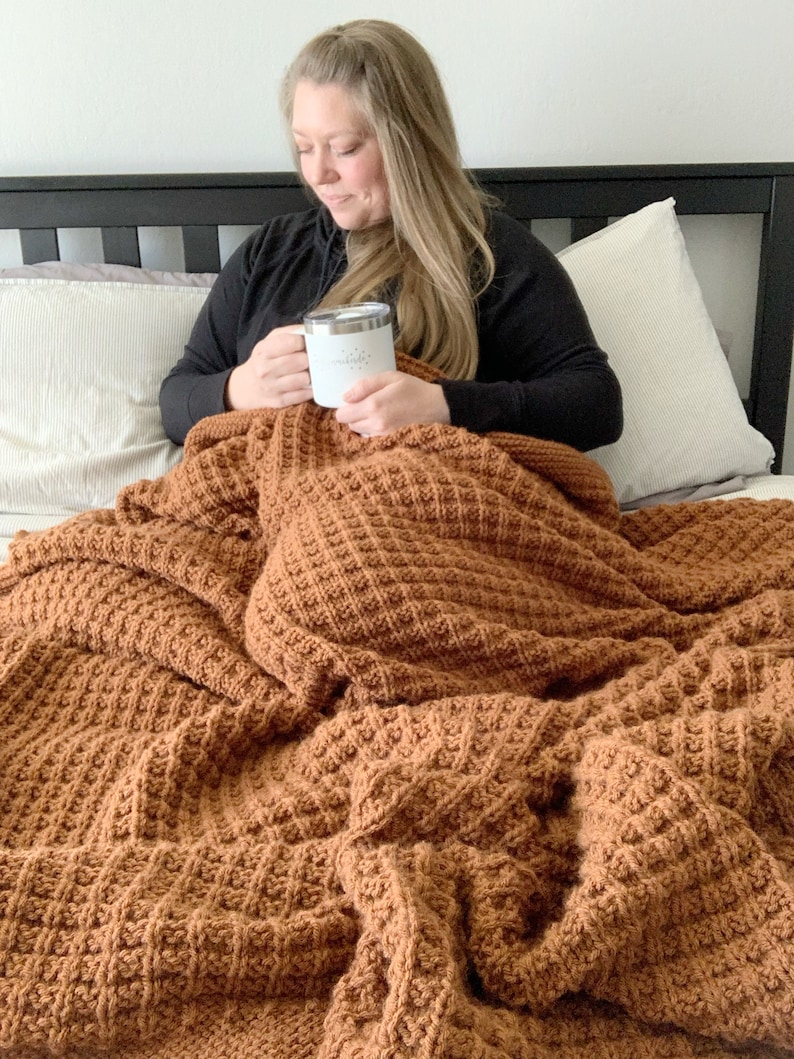May include: A person is sitting on a bed with a brown waffle knit blanket. They are holding a white mug with the word "miracle" on it.