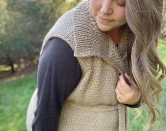 May include: A beige knitted waistcoat with a wide collar and a tie belt. The waistcoat is made of a soft, textured yarn and has a relaxed fit.