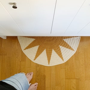 以下が含まれることがあります： ベージュの背景に茶色の星形のデザインが施された、太陽の形をしたかぎ針編みの敷物。敷物は木製の床に置かれています。