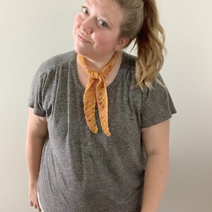 Puede incluir: Una mujer con una camiseta gris y una bufanda de ganchillo naranja y amarilla atada al cuello.