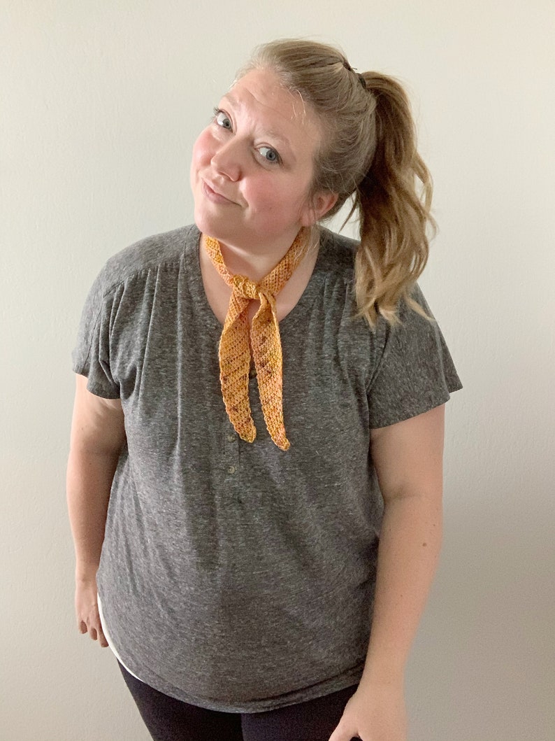 Puede incluir: Una mujer con una camiseta gris y una bufanda de ganchillo naranja y amarilla atada al cuello.