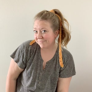 Puede incluir: Una mujer con cabello rubio lleva una camiseta gris y tiene una diadema larga de crochet naranja en el cabello.
