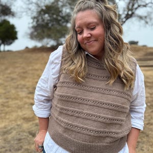 May include: A beige sleeveless knit sweater vest featuring horizontal textured bands and a ribbed neckline, layered over a white long-sleeved button-down shirt and blue denim jeans. The vest has a consistent knit pattern throughout the front.