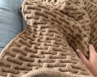 Puede incluir: Una manta tejida de color beige con un patrón texturizado. La manta está doblada, revelando el intrincado diseño. La manta suave y cálida es perfecta para acurrucarse en un día frío. La manta está hecha de un material suave y cálido.
