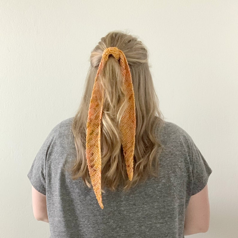 Puede incluir: Una mujer con el pelo largo, ondulado y casta&ntilde;o claro, lleva una camiseta gris con una cinta para el pelo de ganchillo naranja y amarilla.