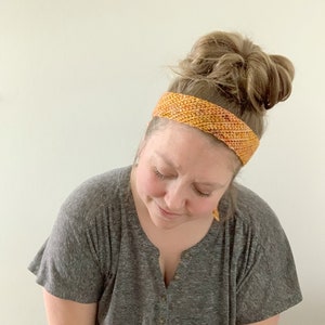 Puede incluir: Una mujer lleva una diadema de ganchillo en tonos naranja y amarillo. La diadema est&aacute; hecha con un patr&oacute;n de punto sencillo y tiene un aspecto ligeramente texturizado.