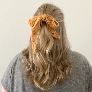 Puede incluir: Un lazo para el pelo de ganchillo en tonos naranja y amarillo, atado en un lazo y usado en el pelo de una mujer.