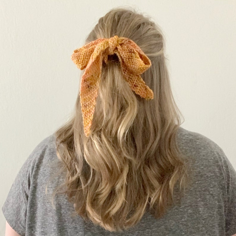 Puede incluir: Un lazo para el pelo de ganchillo en tonos naranja y amarillo, atado en un lazo y usado en el pelo de una mujer.