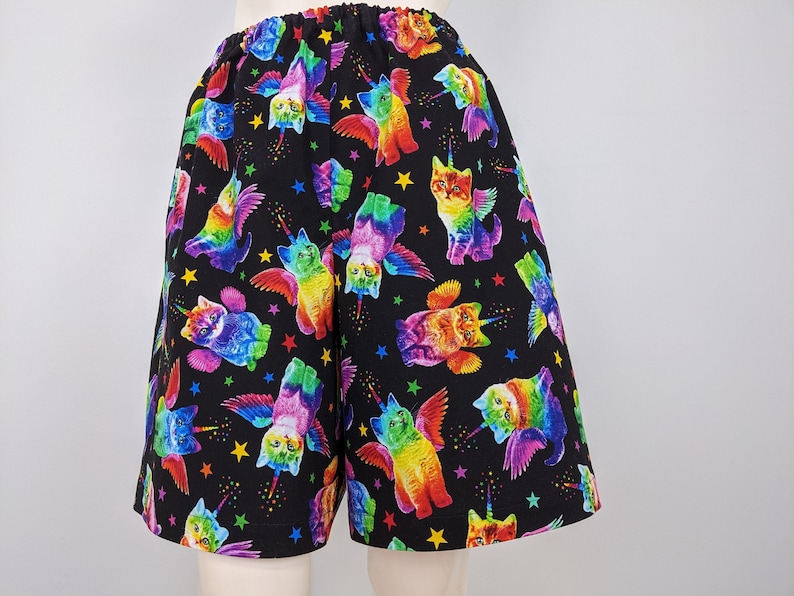 K&ouml;nnte beinhalten: Schwarze Shorts mit elastischem Bund, bedeckt mit einem bunten Muster aus gefl&uuml;gelten Katzen mit Einhorn-H&ouml;rnern und Sternen. Die Katzen haben Regenbogenfarben und sind in verschiedenen Posen.