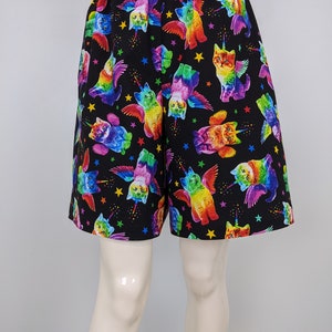 K&ouml;nnte beinhalten: Schwarze Shorts mit einem sich wiederholenden Muster aus regenbogenfarbenen Katzen mit Fl&uuml;geln, Einhorn-H&ouml;rnern und Sternen. Die Shorts haben einen elastischen Bund.