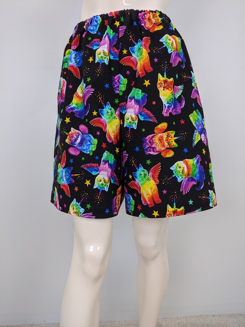 K&ouml;nnte beinhalten: Schwarze Shorts mit einem sich wiederholenden Muster aus regenbogenfarbenen Katzen mit Fl&uuml;geln, Einhorn-H&ouml;rnern und Sternen. Die Shorts haben einen elastischen Bund.