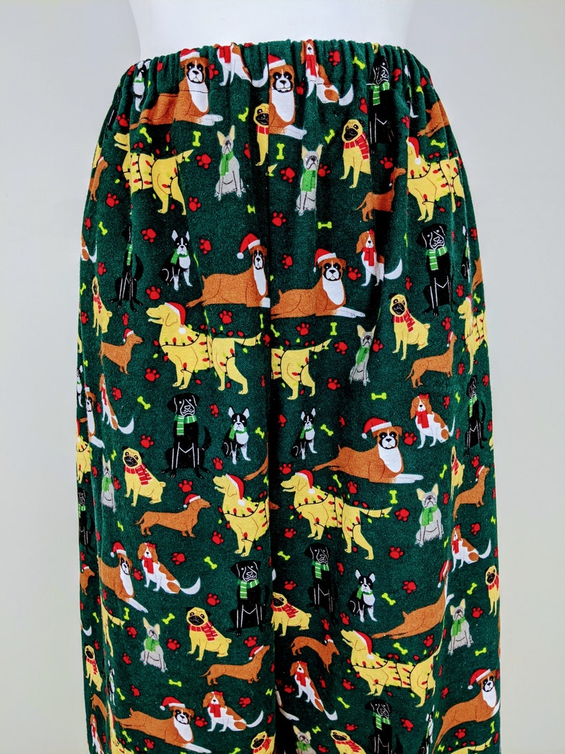 May include: Green pajama pants with an all-over pattern of various dog breeds wearing holiday accessories. The dogs are in brown, yellow, black, and white, with red paw prints and bone accents. The pants have an elastic waistband.