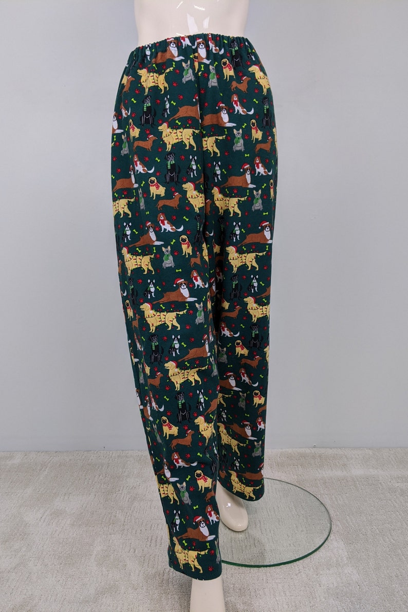 May include: Dark green pajama pants with an all-over pattern of various dog breeds wearing holiday accessories. The dogs are in shades of brown, black, and gold, with red and green accents. The pants have an elastic waistband.