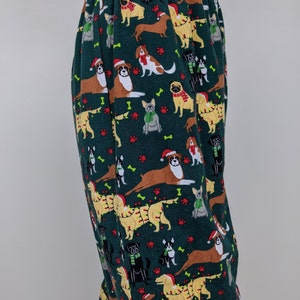 May include: Dark green pajama pants with an all-over pattern of various dog breeds wearing Santa hats, scarves, and Christmas lights. The dogs are in shades of brown, tan, and black, with red paw prints and bone accents. The pants have an elastic waistband.