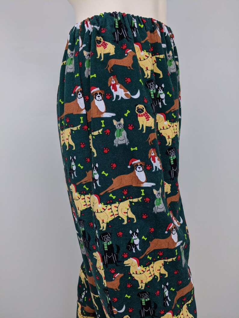 May include: Dark green pajama pants with an all-over pattern of various dog breeds wearing Santa hats, scarves, and Christmas lights. The dogs are in shades of brown, tan, and black, with red paw prints and bone accents. The pants have an elastic waistband.