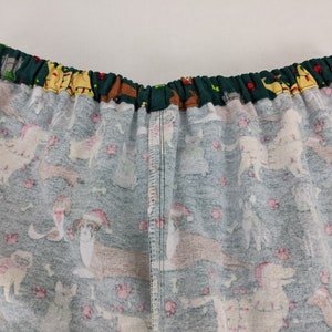 May include: Close-up of pajama bottoms with an elastic waistband. The fabric is a light teal color with a pattern of various dog breeds wearing Santa hats and scarves. Pink paw prints and bones are scattered throughout the design.