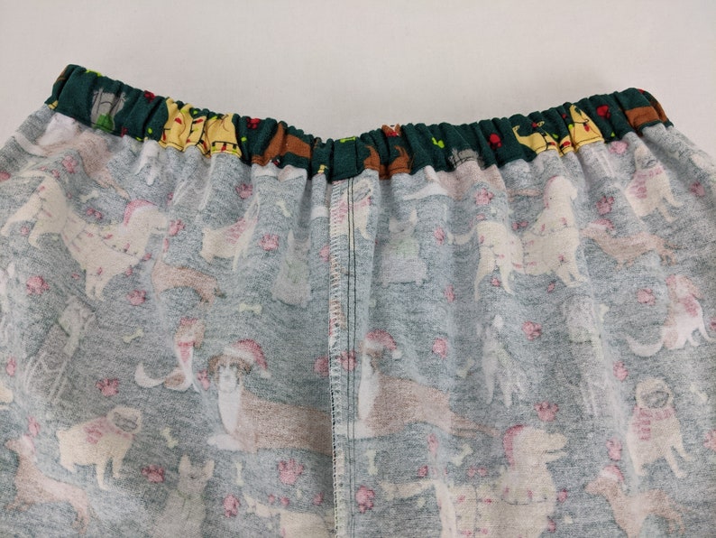 May include: Close-up of pajama bottoms with an elastic waistband. The fabric is a light teal color with a pattern of various dog breeds wearing Santa hats and scarves. Pink paw prints and bones are scattered throughout the design.