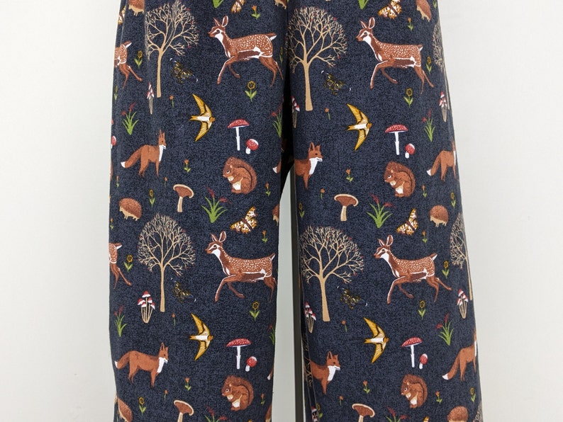 May include: Dark blue fabric with a repeating pattern of woodland creatures and elements. The design includes deer, foxes, squirrels, mushrooms, butterflies, birds, and trees. The fabric is likely used for clothing or home decor.