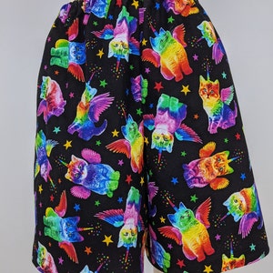 K&ouml;nnte beinhalten: Schwarze Shorts mit elastischem Bund, bedeckt mit einem farbenfrohen Muster aus gefl&uuml;gelten Katzen-Einh&ouml;rnern und Sternen. Die Katzen-Einh&ouml;rner sind in verschiedenen Posen und Regenbogenfarben dargestellt. Die Shorts sind aus weichem Stoff.