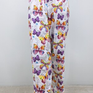 Puede incluir: Pantalones blancos con estampado de mariposas de colores. Las mariposas son moradas, amarillas, naranjas y rosas.