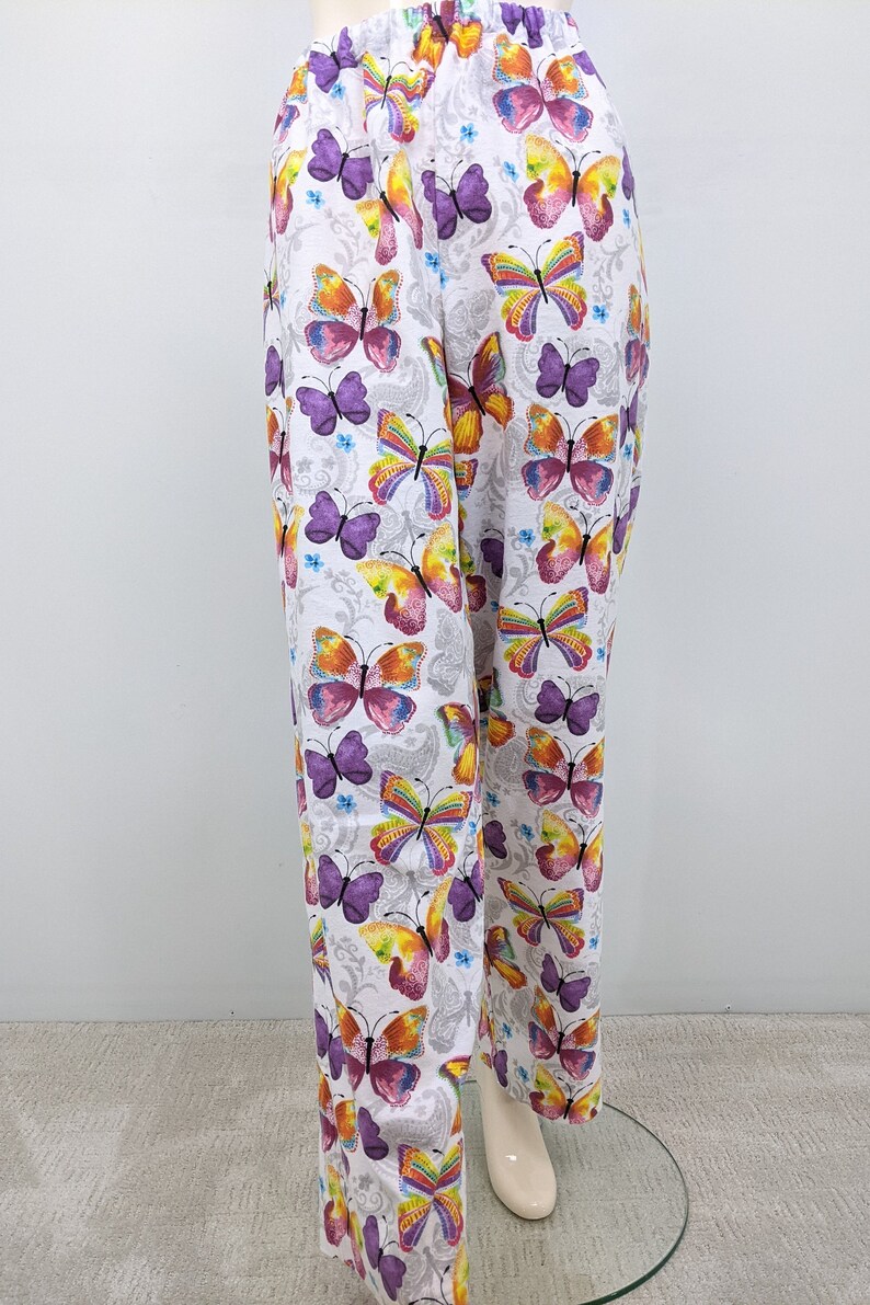 Puede incluir: Pantalones blancos con estampado de mariposas de colores. Las mariposas son moradas, amarillas, naranjas y rosas.