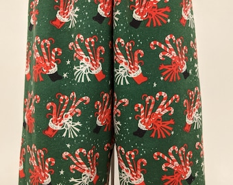 Puede incluir: Pantalones de pijama verdes con un estampado repetido de bastones de caramelo rojos y blancos, medias rojas y lazos blancos. El fondo tiene pequeñas estrellas blancas. Los pantalones son de tela suave.