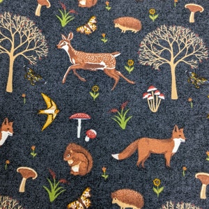 May include: Dark gray fabric with a repeating pattern of woodland creatures and plants. The design includes foxes, deer, squirrels, mushrooms, butterflies, trees, and birds. The illustrations are in shades of brown, yellow, and white.