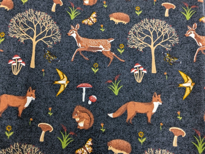 May include: Dark gray fabric with a repeating pattern of woodland creatures and plants. The design includes foxes, deer, squirrels, mushrooms, butterflies, trees, and birds. The illustrations are in shades of brown, yellow, and white.