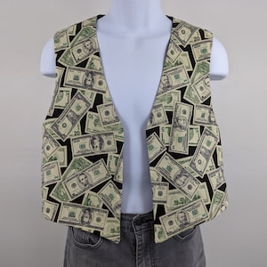 May include: A sleeveless vest featuring a repeating pattern of U.S. dollar bills in shades of green, black, and cream. The vest is open in the front and appears to be made of a lightweight fabric. The background is a solid white.