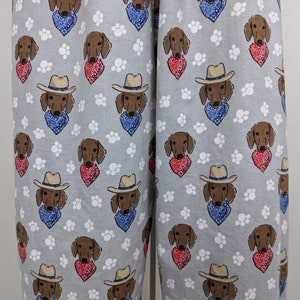 Könnte beinhalten: Hellgraue Schlafanzughose mit einem sich wiederholenden Muster aus braunen Dackeln mit Cowboyhüten und Bandanas. Einige Hunde haben blaue Bandanas, andere rote. Weiße Pfotenabdrücke sind über den Stoff verstreut.