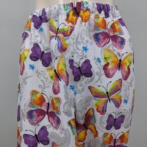 Puede incluir: Pantalones blancos con estampado de mariposas de colores. Las mariposas son de color morado, naranja, amarillo y rosa.
