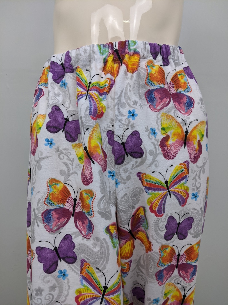 Puede incluir: Pantalones blancos con estampado de mariposas de colores. Las mariposas son de color morado, naranja, amarillo y rosa.