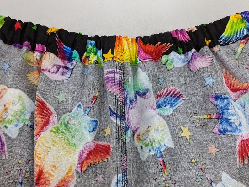K&ouml;nnte beinhalten: Nahaufnahme eines Stoffes mit einem sich wiederholenden Muster aus regenbogenfarbenen Katzen-Einh&ouml;rnern mit Fl&uuml;geln und Sternen auf grauem Hintergrund. Der obere Rand ist schwarz mit Regenbogensternen. Der Stoff wird wahrscheinlich f&uuml;r Kleidung oder Accessoires verwendet.