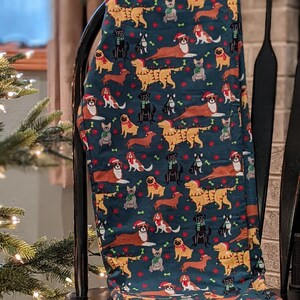 May include: Dark teal leggings featuring a repeating pattern of various dog breeds wearing Santa hats and scarves. The dogs are in shades of brown, tan, black, and white, with red paw prints and bones scattered throughout the design. The leggings are draped over a black chair.
