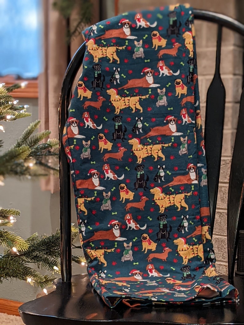 May include: Dark teal leggings featuring a repeating pattern of various dog breeds wearing Santa hats and scarves. The dogs are in shades of brown, tan, black, and white, with red paw prints and bones scattered throughout the design. The leggings are draped over a black chair.