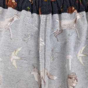 May include: Close-up of fabric with a woodland animal print. The fabric features deer, foxes, birds, mushrooms, and other forest elements in shades of gray, brown, and white. The top edge is a dark gray elastic waistband.