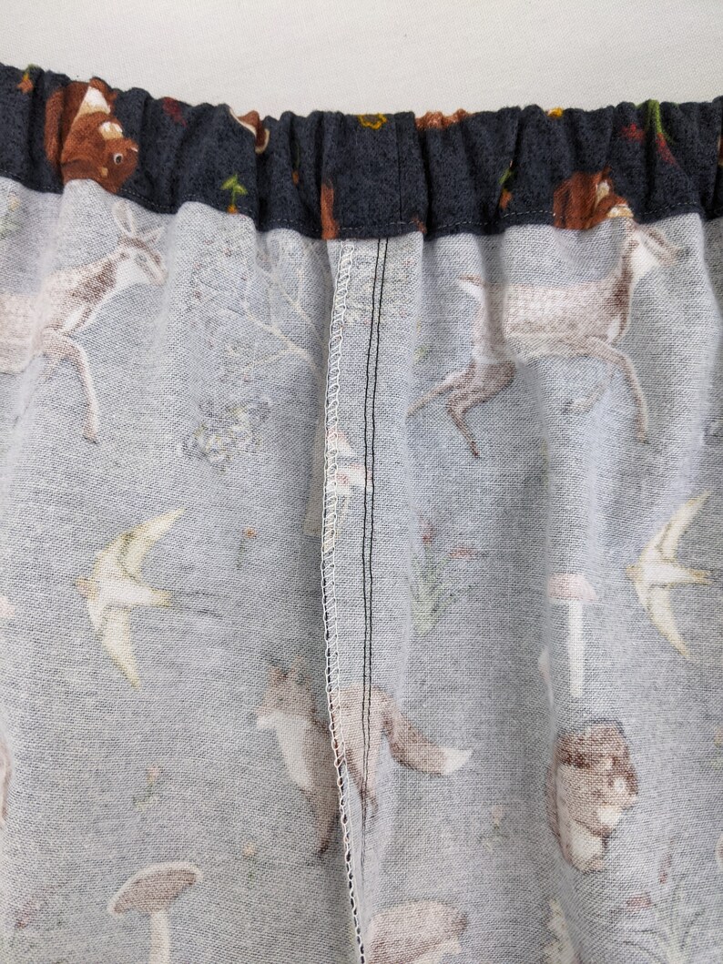 May include: Close-up of fabric with a woodland animal print. The fabric features deer, foxes, birds, mushrooms, and other forest elements in shades of gray, brown, and white. The top edge is a dark gray elastic waistband.