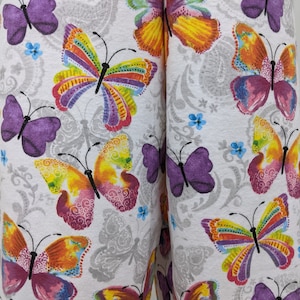 Puede incluir: Tela blanca con un patr&oacute;n repetido de mariposas coloridas. Las mariposas tienen acentos morados, amarillos, naranjas, rosados y verdes.