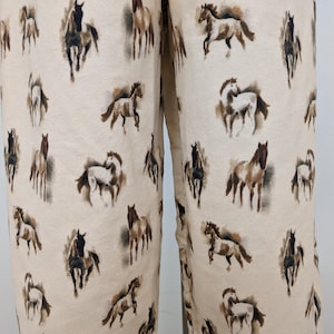 Könnte beinhalten: Eine cremefarbene Hose mit einem Allover-Druck von braunen, schwarzen und weißen Pferden in verschiedenen Posen. Die Pferde sind in einem malerischen Stil dargestellt und in einem sich wiederholenden Muster über den gesamten Stoff verteilt.