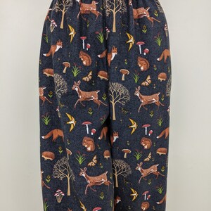 May include: Dark blue pajama pants with an elastic waistband. The fabric features a repeating pattern of woodland creatures, including deer, foxes, squirrels, hedgehogs, butterflies, and mushrooms. The trees and mushrooms are brown and red.