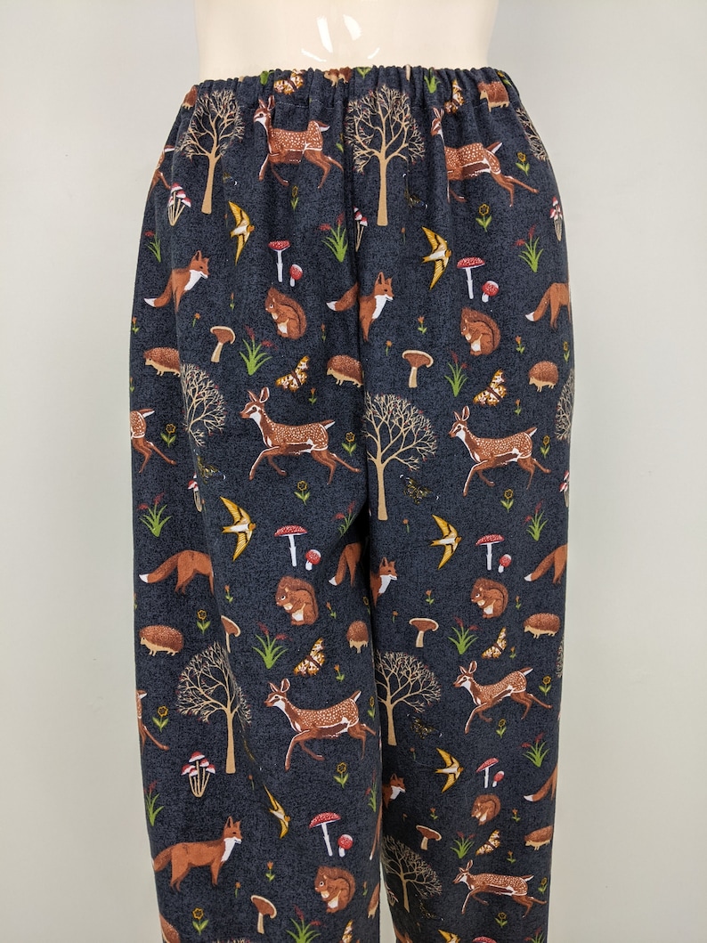 May include: Dark blue pajama pants with an elastic waistband. The fabric features a repeating pattern of woodland creatures, including deer, foxes, squirrels, hedgehogs, butterflies, and mushrooms. The trees and mushrooms are brown and red.