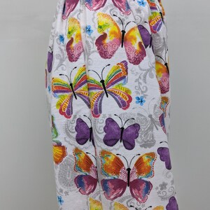 Puede incluir: Pantalones blancos con estampado de mariposas de colores. Las mariposas son en tonos de rosa, morado, amarillo, naranja y verde. El fondo es blanco con un patr&oacute;n floral gris.