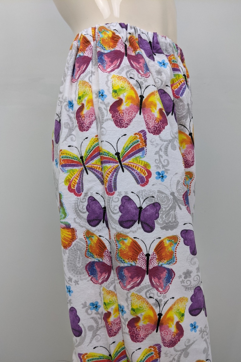 Puede incluir: Pantalones blancos con estampado de mariposas de colores. Las mariposas son en tonos de rosa, morado, amarillo, naranja y verde. El fondo es blanco con un patr&oacute;n floral gris.