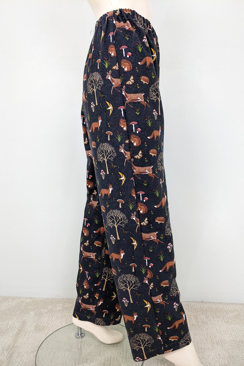 May include: Dark navy blue pajama pants with an all-over woodland print. The pattern includes brown foxes, deer, mushrooms, trees, butterflies, and hedgehogs. The pants have an elastic waistband.