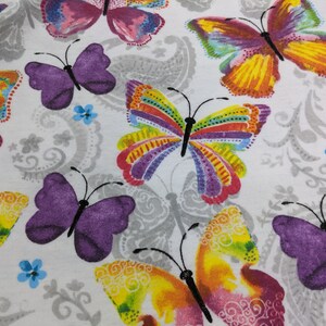 Puede incluir: Tela blanca con un patr&oacute;n repetitivo de mariposas de colores. Las mariposas tienen alas moradas, rosas, amarillas, naranjas, rojas, verdes y azules.
