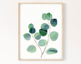 Puede incluir: Pintura acuarela de una rama de eucalipto con hojas verdes sobre un fondo blanco. La pintura está enmarcada en un marco de madera claro.