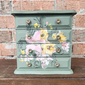 May include: A small green wooden jewelry box with four drawers. The drawers are decorated with a floral print in shades of pink, yellow, and white. The box has gold-toned metal knobs.