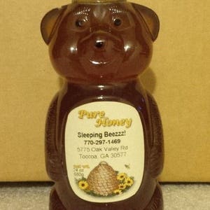 May include: A brown bear-shaped bottle of honey with a yellow lid. The label reads "Pure Honey", "Sleeping Beezzz!", "770-297-1469", "5775 Oak Valley Rd", "Toccoa, GA 30577", "24 oz", and "680g".